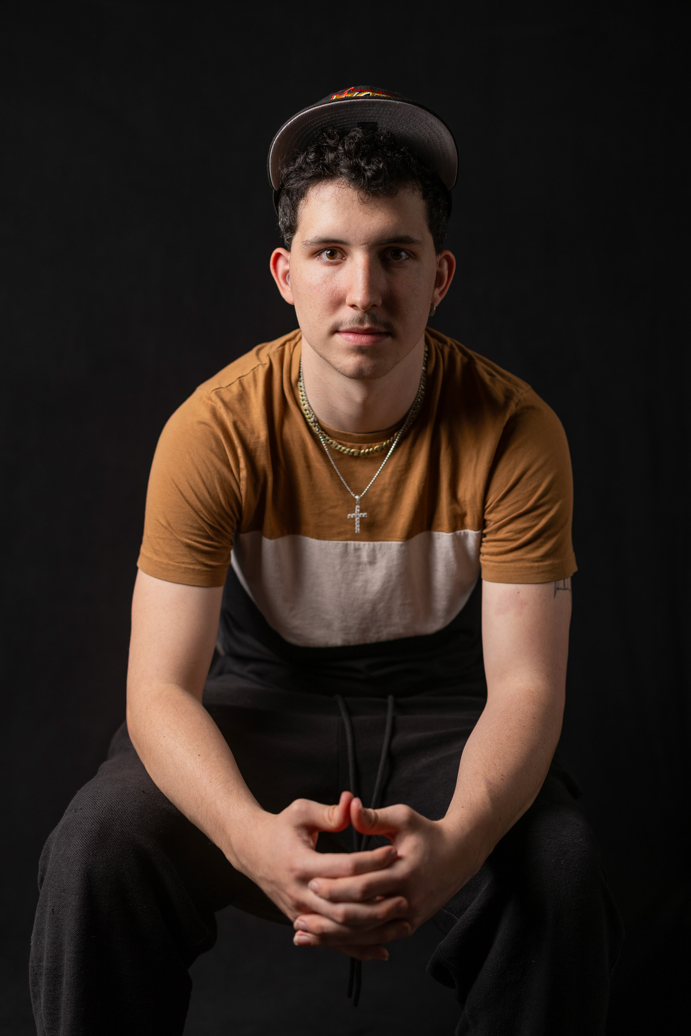 A young man is seated in a chair with his elbows resting on his thighs as he leans forward towards the camera. He looks straight ahead with dark curly hair and wears a baseball cap. He is illuminated by light and there is a dark black background behind him. 