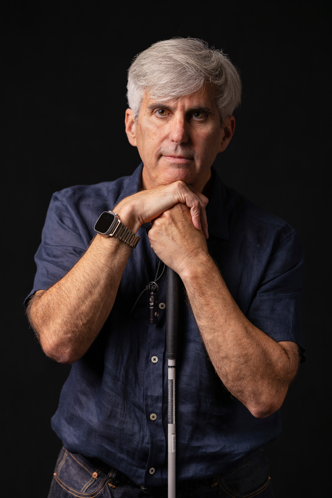 A man looks straight ahead and rests his chin and both hands on his mobility cane. He has light gray hair and he is illuminated by light with shadow on one half of his face. There is a dark black background behind him.