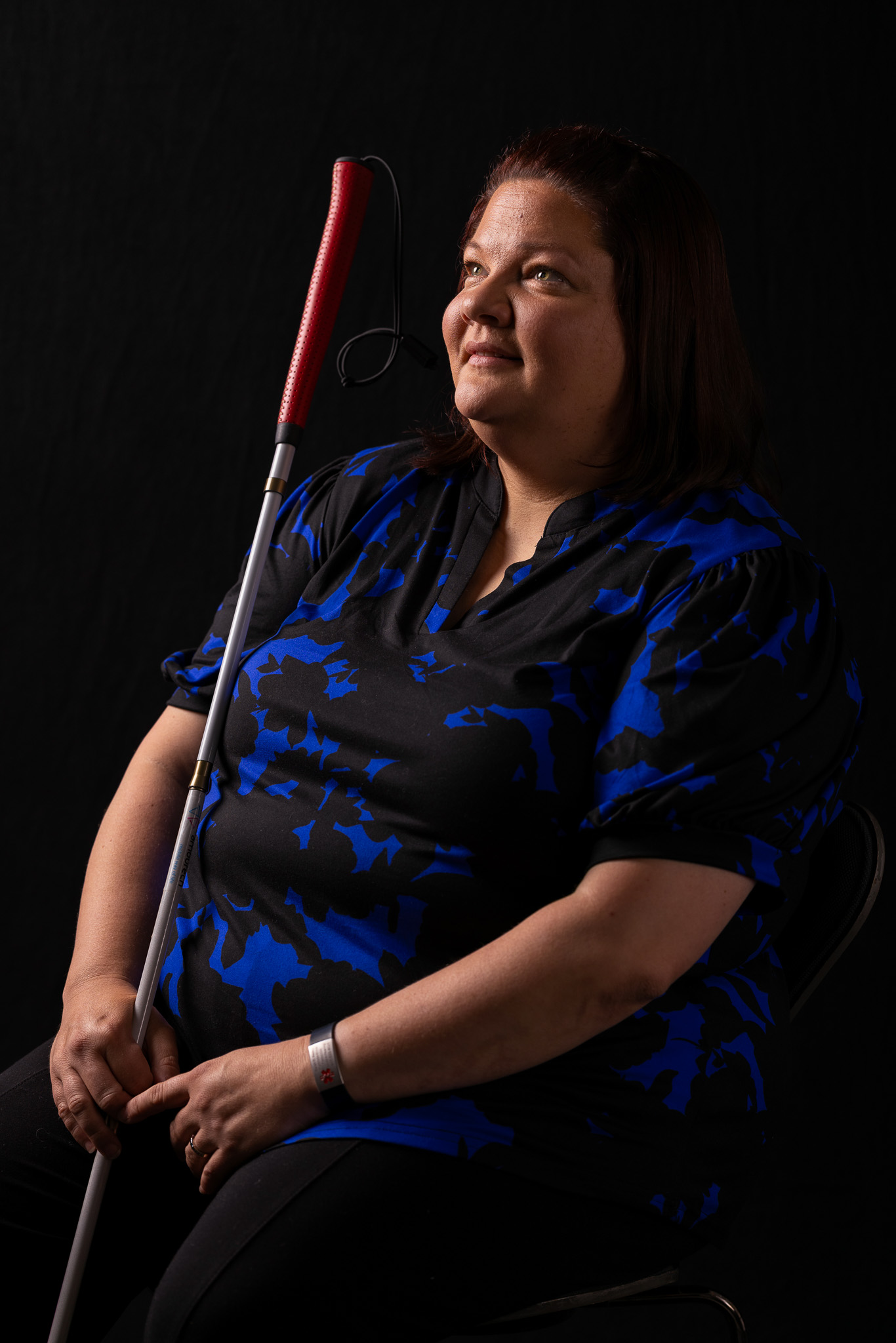 A woman is seated at an angle looking straight ahead. She holds a cane in one hand and her face is illuminated by light. There is a dark black background behind her. She has a soft smile on her face and wears a blue short sleeve shirt.