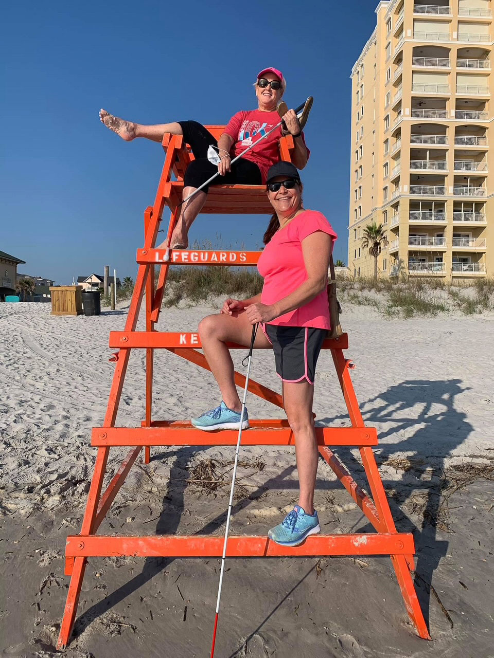 two woman climb a lifeguard stand on a sandy beach and they are holding canes.