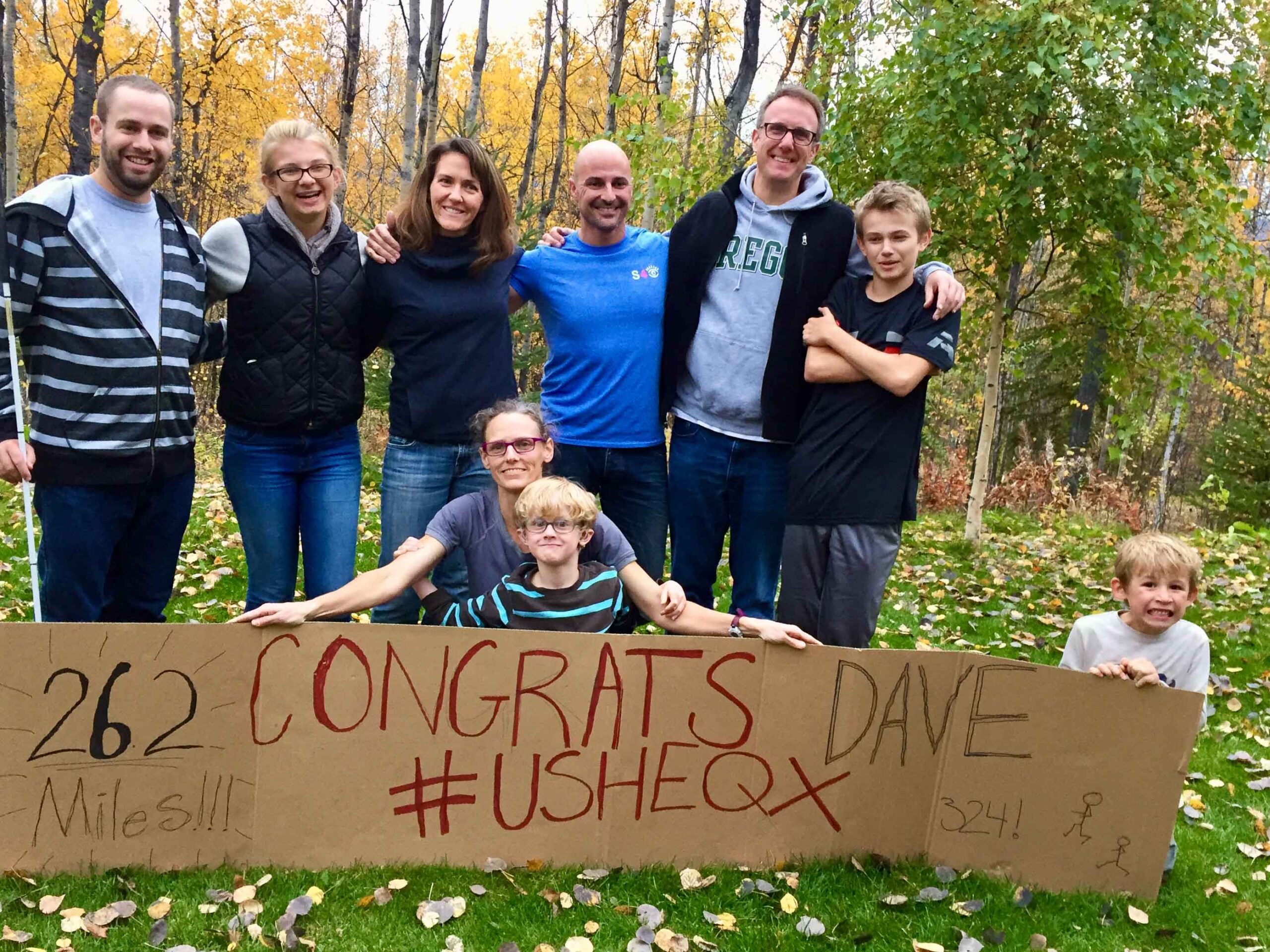 A group photo with 8 people holding a large marathon sign that says "26.2 Marathon Congrats Dave #USHEQX"