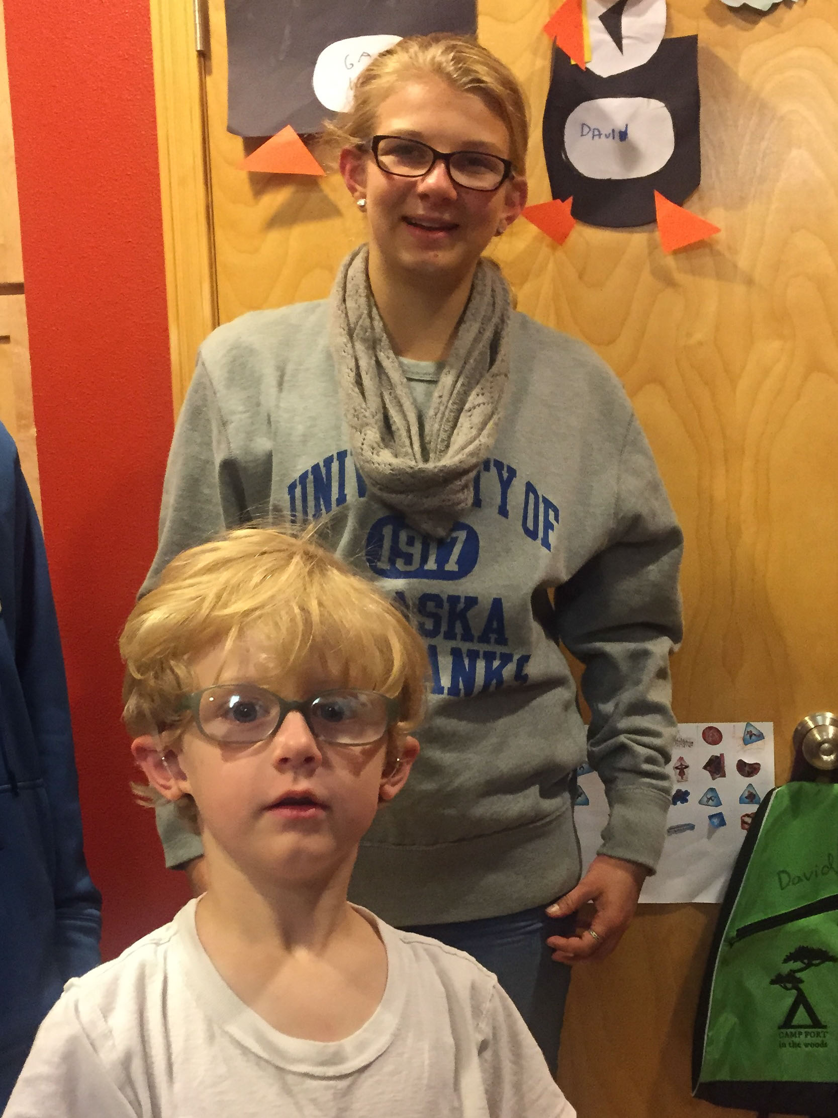 A photo of a young woman, Bella, and a little boy, Galen standing together in what appears to be a classroom. There is a door behind them with cut paper artwork. Both Bella and Galen wear glasses and look ahead at the camera. 