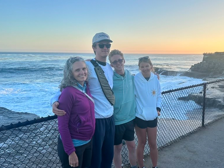 A group photo of people standing outside in front of the water during a sunset. There are 4 people in the photo, they are all smiling and looking a the camera.