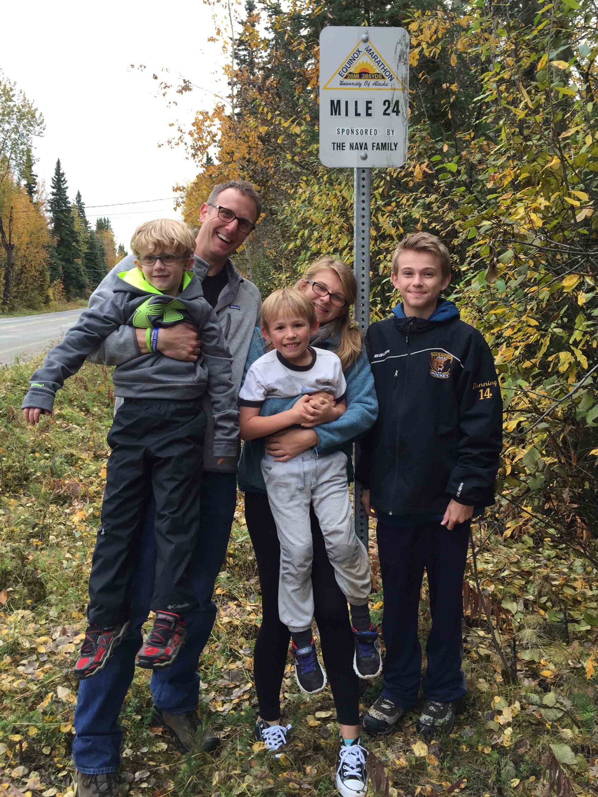A group photo of 5 people standing on a hiking trail. Two people standing in the back of the group are holding two of the younger kids in front of them.