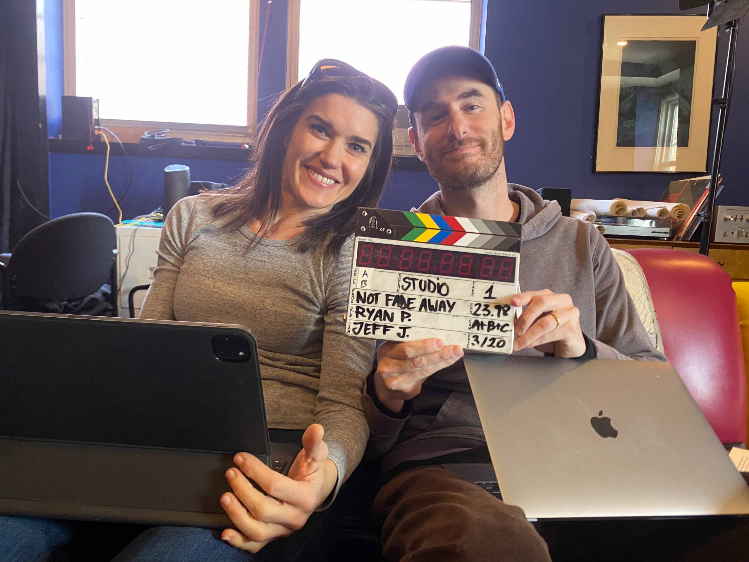 Rebecca Alexander sits next to Ryan Porush on a couch and they hold up a clapperboard for Not Fade Away