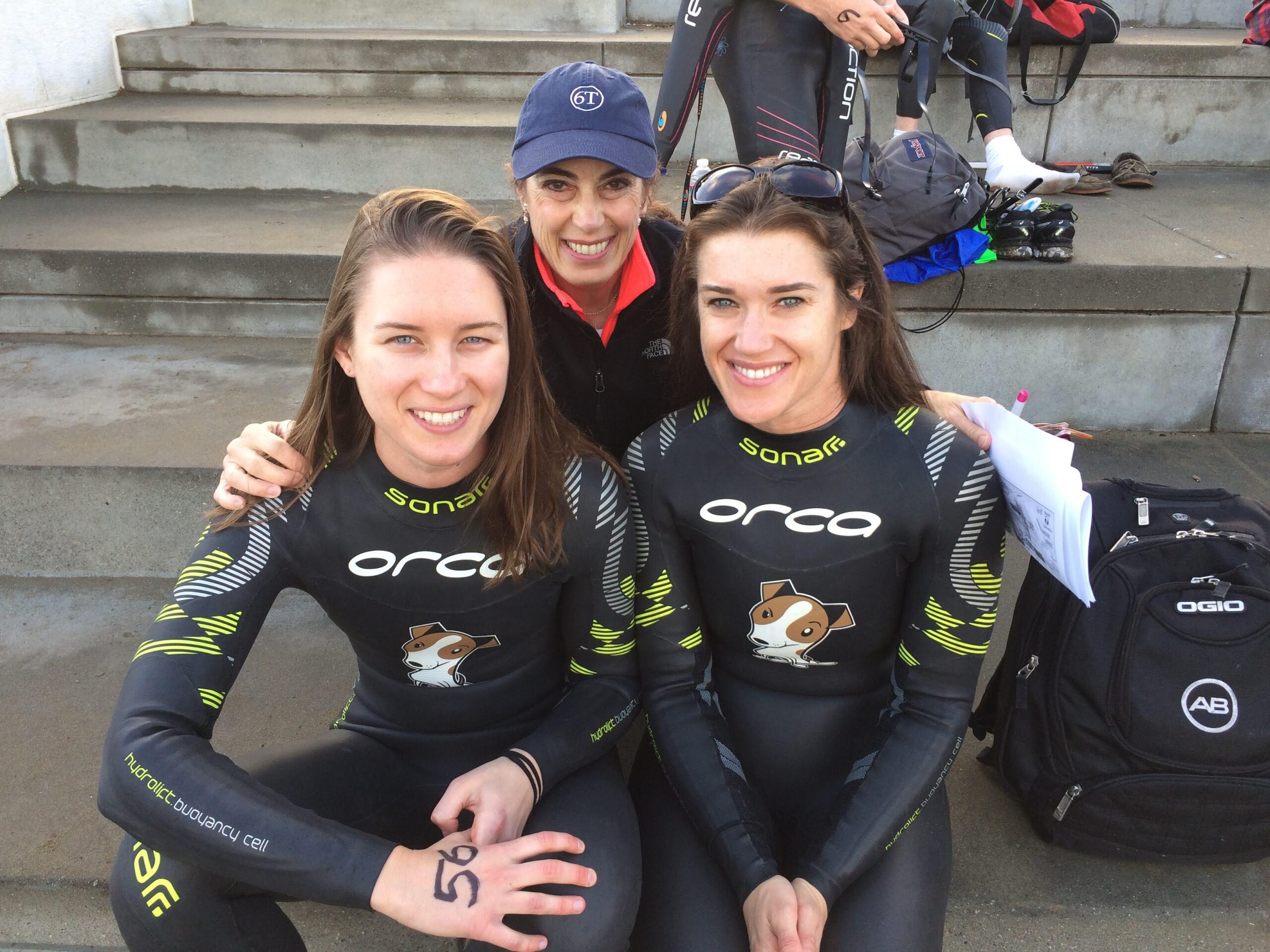 Three women sit together on outdoor concrete steps and smile for a photo. Two of the women, Rebecca and her friend Caroline, are wearing black wetsuits.