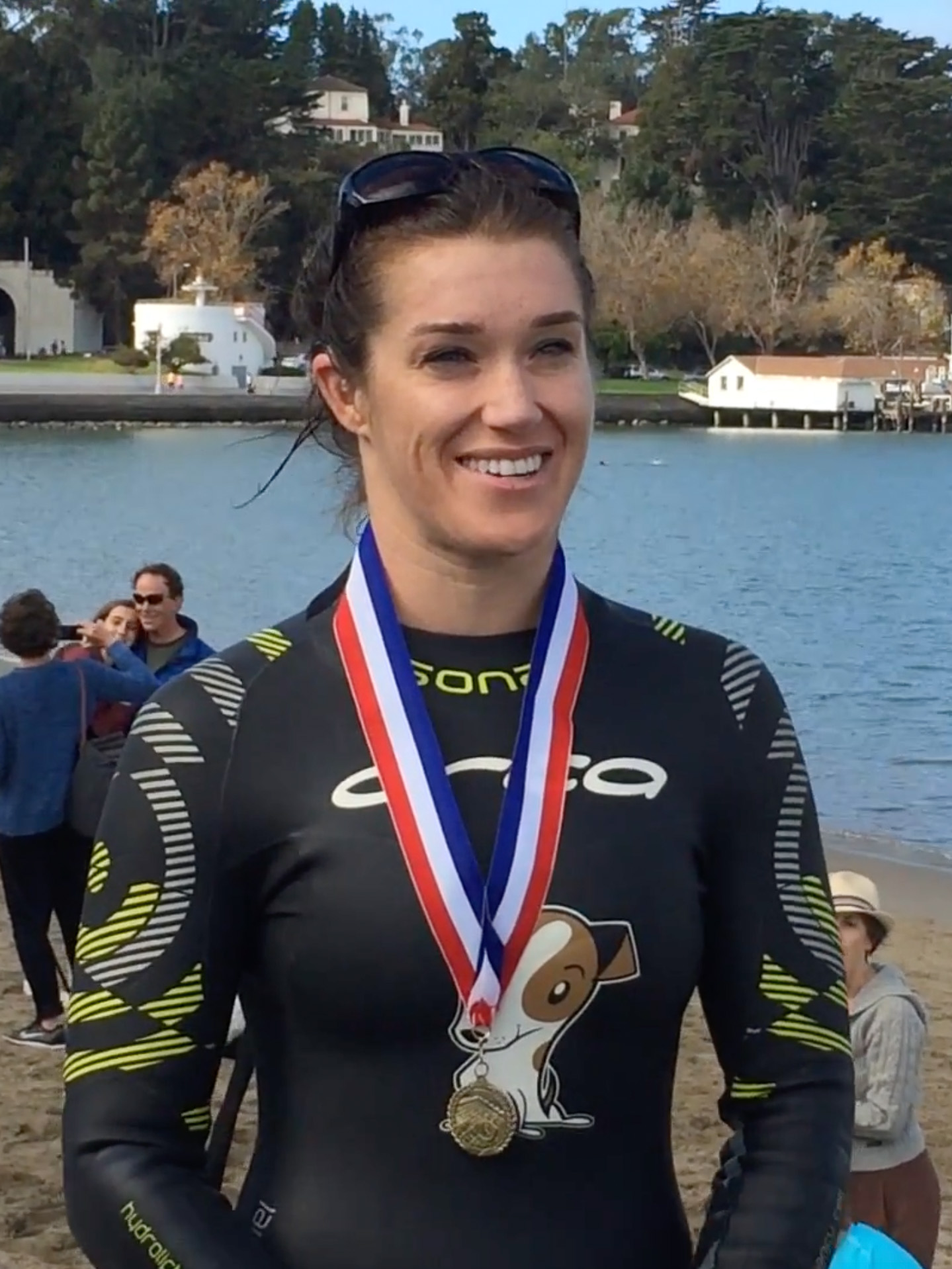 A photo of a woman wearing a wetsuit and a metal. She is smiling and there is water behind her. 