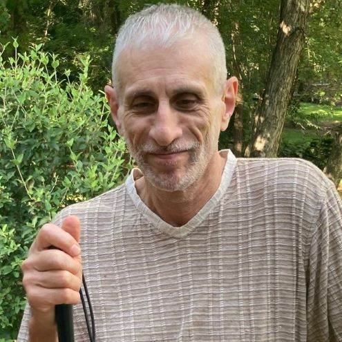 Carlos in 2026 holding his white cane standing on his back deck at his home in Camden